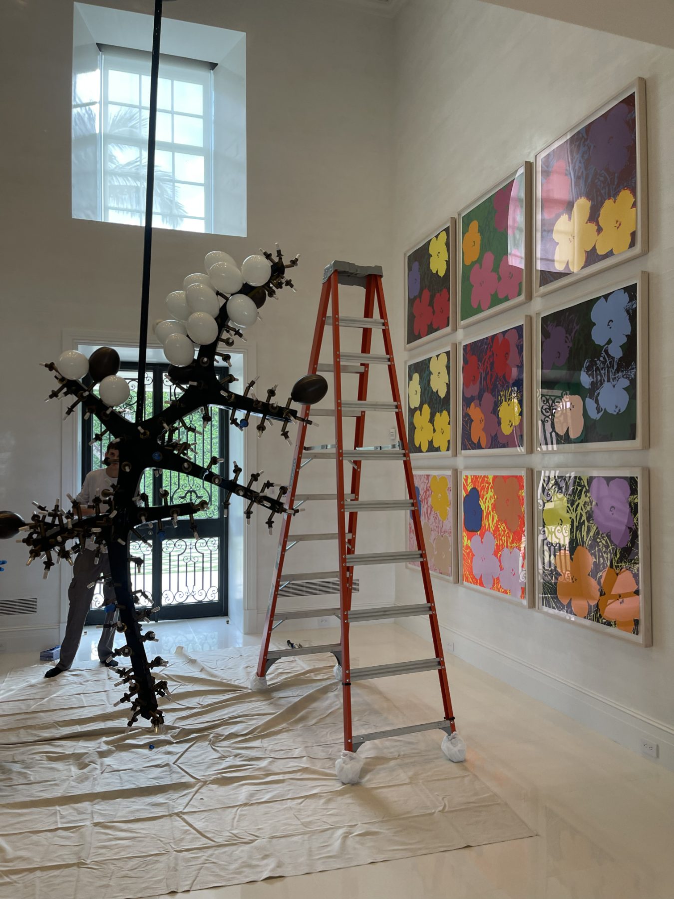 Installation underway in a designer residence: a grid of framed prints hung on the wall beside a sculptural light fixture, floors covered with drop cloths